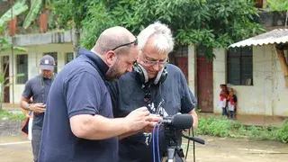Two men adjusting audio equipment on a tripod outdoors, surrounded by greenery and buildings in the background.